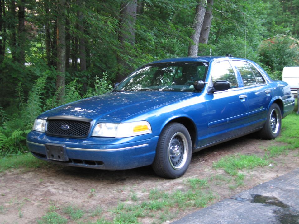 Blue 2001 Ford Crown Victoria parked on dirt and grass with trees in background