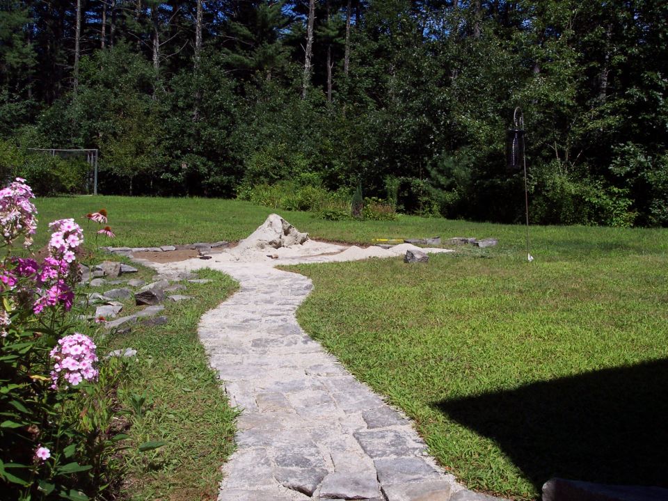 stone pathway leading through a grassy backyard with a sand pit and rocks, surrounded by trees
