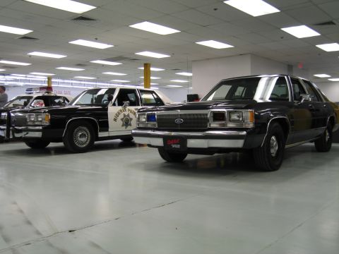 2001 Ford Crown Victoria - Two 1980s-90s Ford LTD Crown Victoria police sedans displayed on a polished shop floor: a black unmarked car in front with a blue grille-mo…
