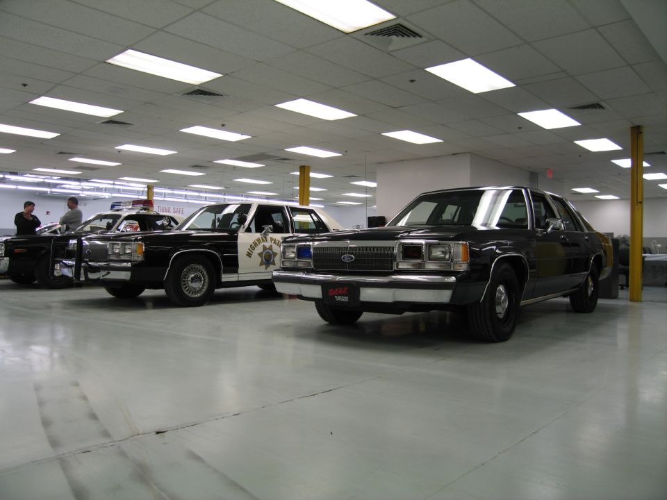 Two police cars parked indoors, one black and one black and white. The black and white car has 'HIGHWAY PATROL' on its door