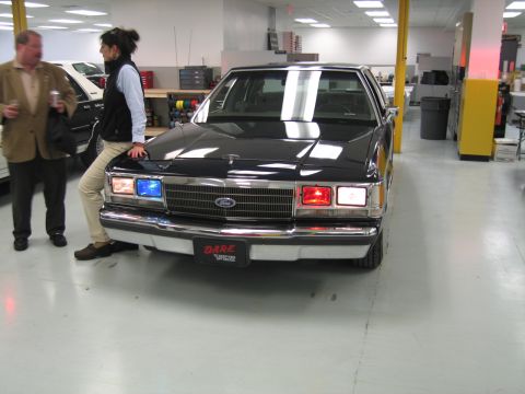 2001 Ford Crown Victoria - A dark 2001 Ford Crown Victoria, equipped with a DARE license plate and emergency lights, is shown from the front.