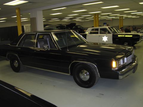 2001 Ford Crown Victoria - A black sedan is parked indoors next to a white and black police car with "DEPUTY SHERIFF" text.