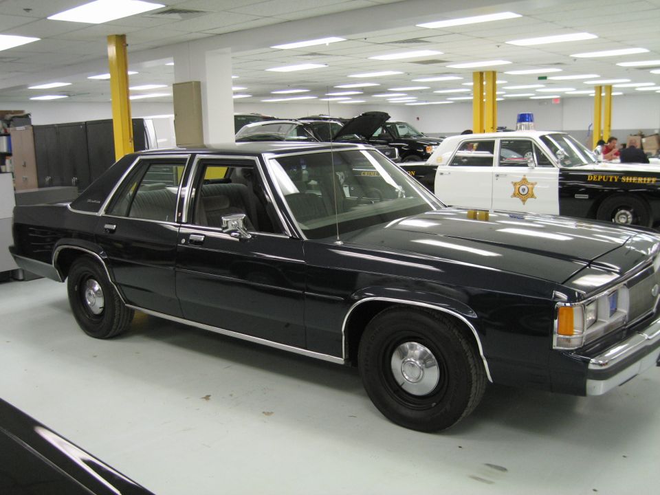 Black sedan, 3/4 front view. Another police car is visible in the background
