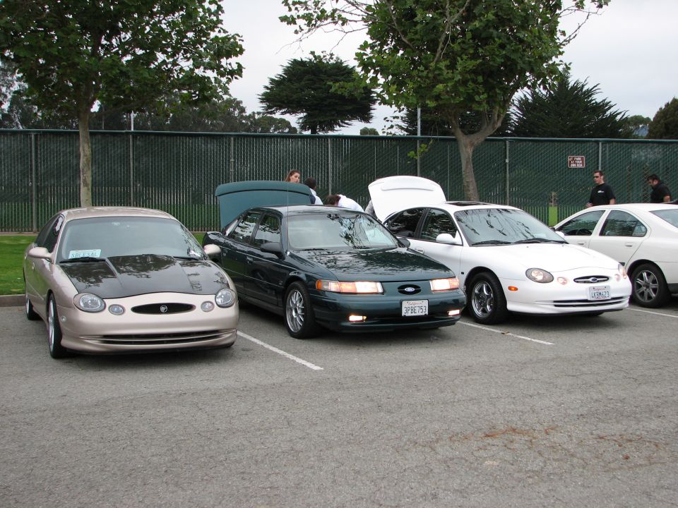 Three sedans parked in a lot, one tan with a black hood, one dark green, and one white