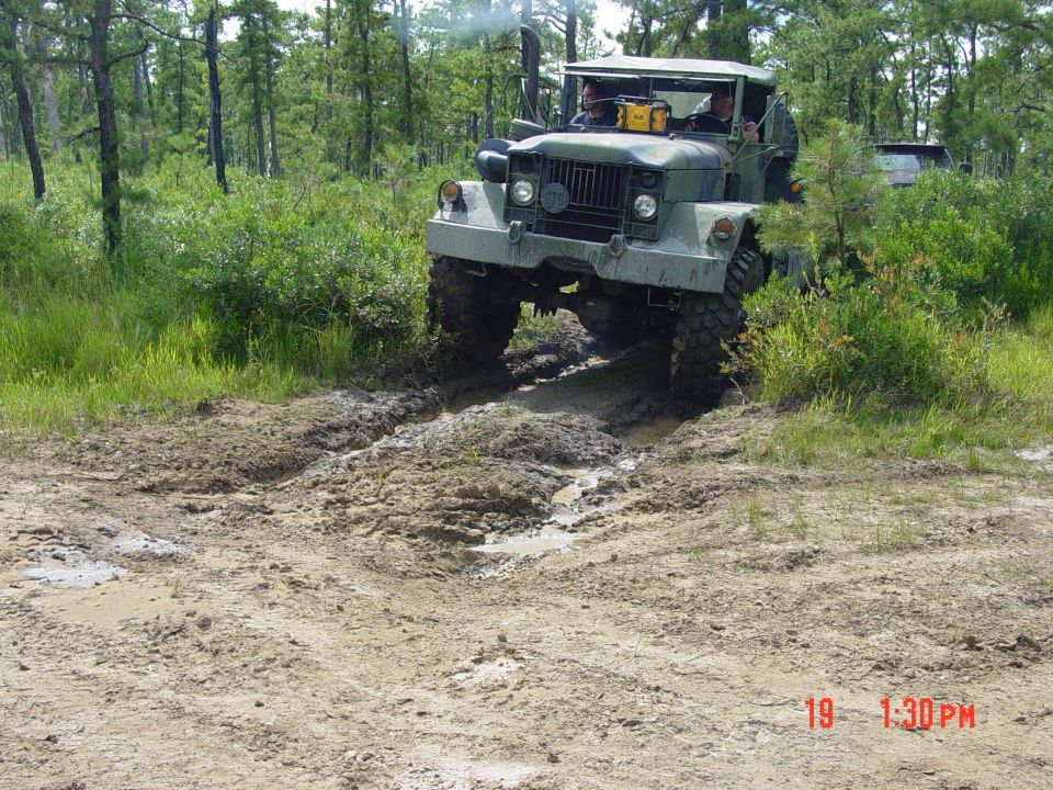 Military truck with large tires driving through a muddy wooded area. Another vehicle is partially visible behind it
