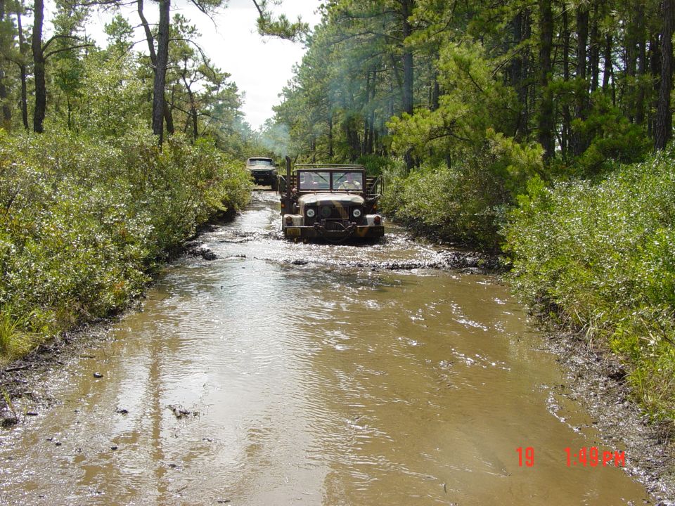 Two old military trucks driving through a muddy, wooded trail