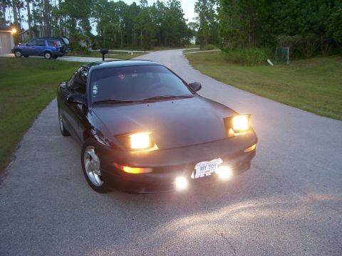 1994 Ford Probe - A black coupe with its distinctive pop-up headlights illuminated is parked on a paved road.