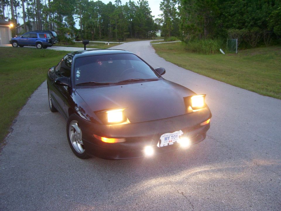 Black coupe with pop-up headlights on a road. License plate says MY TOY
