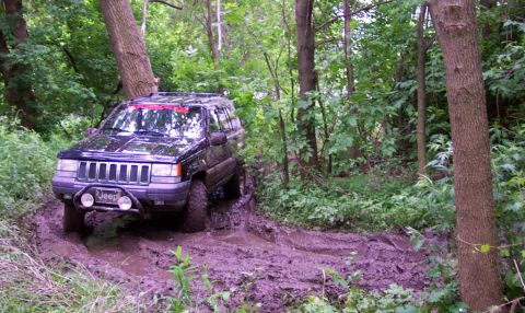 1978 Ford Bronco - A black Jeep Grand Cherokee tackles a challenging muddy path through a wooded area.