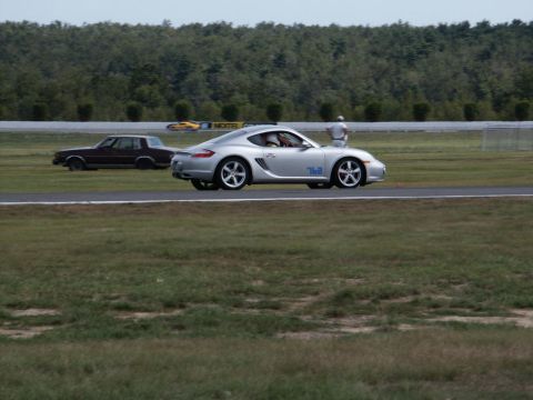 2001 Mercury Grand Marquis - A silver Porsche Cayman and a dark sedan are on a race track with trees in the background.