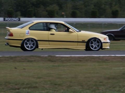 2001 Mercury Grand Marquis - A yellow BMW coupe is seen on a race track, with a driver wearing a helmet. A sign that says POCONO is visible in the background.