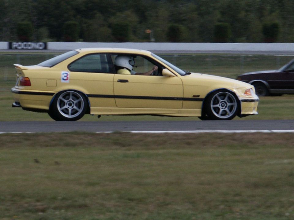 Yellow BMW coupe with racing number 3 on door, driver wearing helmet, on a race track. Another car is visible in the background