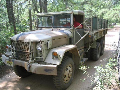 1990 Ford Bronco - This military truck is driving on a dirt road through a forest. The vehicle has a camouflage paint scheme.