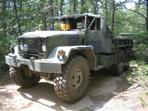 1990 Ford Bronco - A military truck with a flatbed is driven off-road through a wooded area.