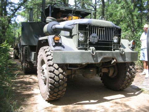 1990 Ford Bronco - A large, olive-drab military truck with a prominent grille is shown on a wooded dirt path.