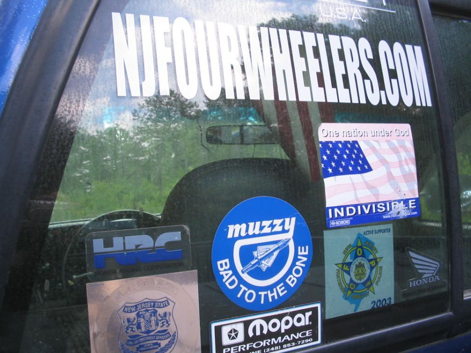 Blue vehicle interior seen through window covered in stickers including NJFOURWHEELERS.COM, Muzzy, Mopar, and an American flag