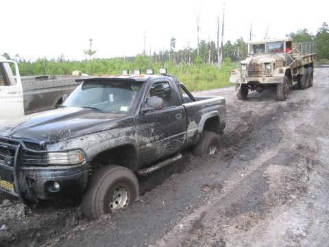 1990 Ford Bronco - A black pickup truck is stuck in deep mud and being towed by a vintage military truck on a dirt road.