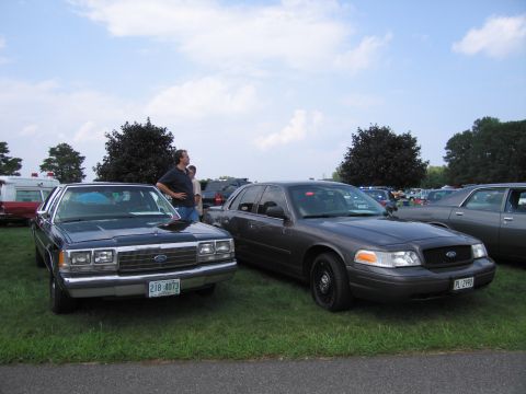 2001 Ford Crown Victoria - Two Ford Crown Victorias are parked on a grassy field under a partly cloudy sky.