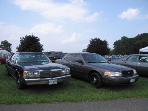 2001 Ford Crown Victoria - Two Ford Crown Victoria police cars are parked side-by-side on a grassy field under a partly cloudy sky.