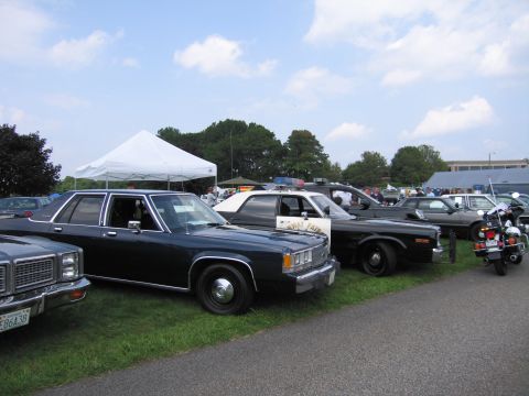 2001 Ford Crown Victoria - Several older model police cars and a motorcycle are parked on a grassy area under a tent.