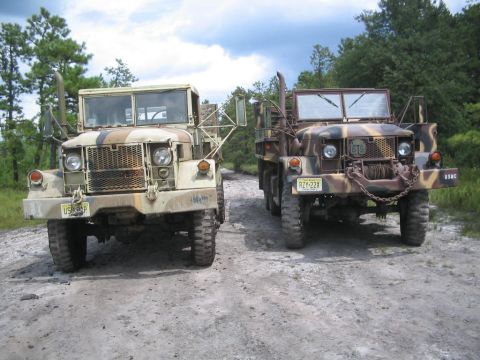 1990 Ford Bronco - Two military trucks are parked side-by-side on a dirt road with trees in the background.