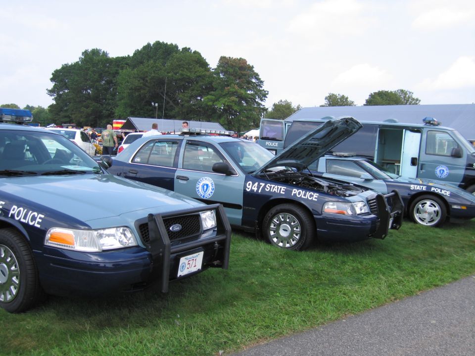 Two blue and grey police cars parked on grass, one with hood open. The front car has a push bumper and license plate 571