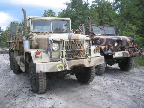 1990 Ford Bronco - Two military trucks are parked on a dirt road with trees in the background.