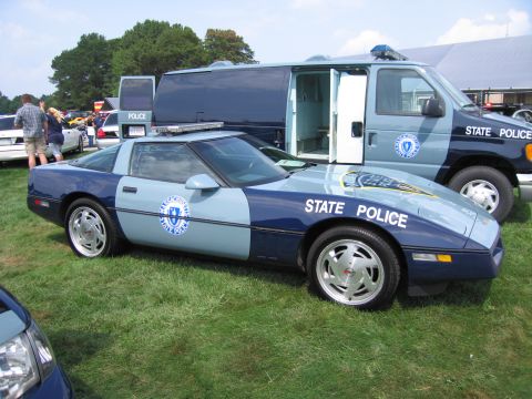2001 Ford Crown Victoria - A police Corvette and van are parked on a grassy field under a partly cloudy sky.