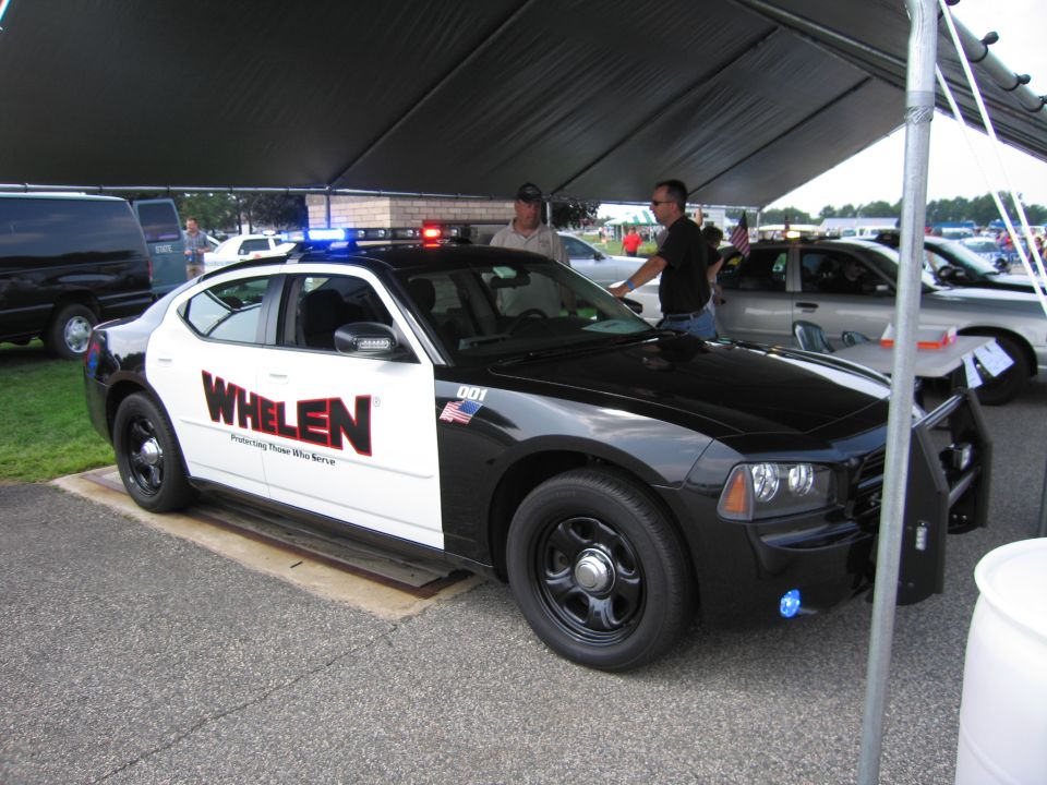 Black and white police car with Whelen logo on the side, parked under a tent. Two men stand near the car