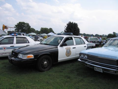2001 Ford Crown Victoria - Two police cars and a light blue car are parked on a grassy field under a cloudy sky.