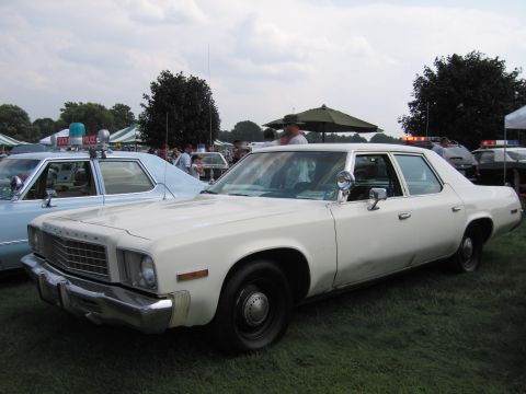 2001 Ford Crown Victoria - A white sedan, possibly a Plymouth, is parked on grass next to a light blue police car.