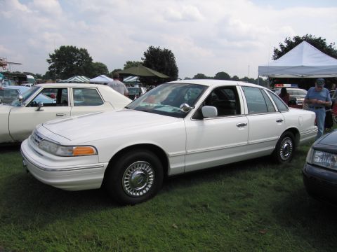 2001 Ford Crown Victoria - A white 2001 Ford Crown Victoria is parked on grass at an outdoor event, with other vehicles and tents in the background.