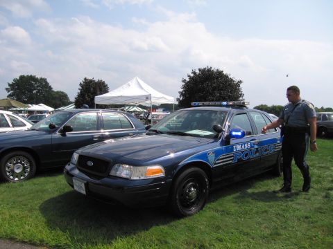 2001 Ford Crown Victoria - A police officer stands next to a UMass Police car, with another vehicle parked nearby on a grassy field under a partly cloudy sky.