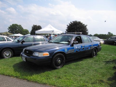 2001 Ford Crown Victoria - A blue 2001 Ford Crown Victoria police car is parked on a grassy field with other vehicles and tents in the background.