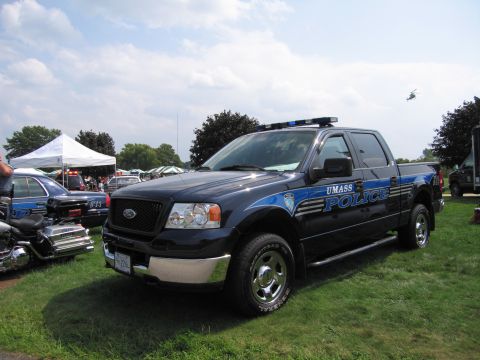2001 Ford Crown Victoria - A dark blue UMASS POLICE truck is parked on grass, with other vehicles and tents in the background. A helicopter flies in the sky.