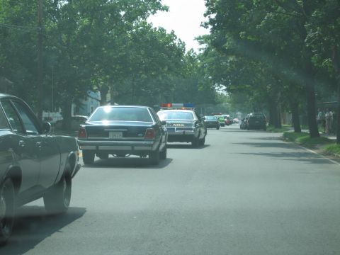 2001 Ford Crown Victoria - A line of vehicles, including marked police cars, proceeds down a suburban street on a sunny day.