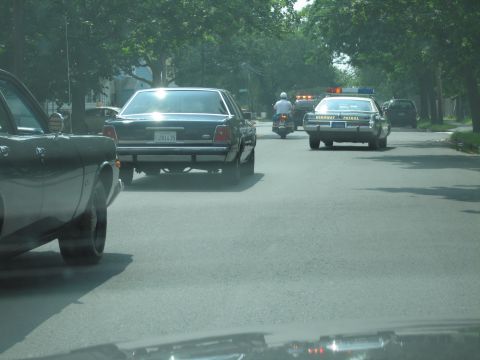 2001 Ford Crown Victoria - A dark sedan with license plate 2R1L25 is seen from behind, with a police car and motorcycle in the distance.