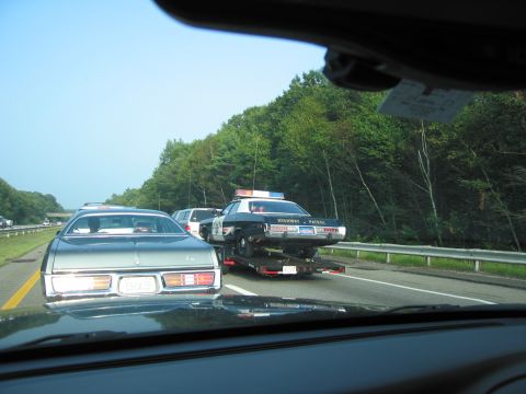 2001 Ford Crown Victoria - A police car is transported on a trailer on the highway, its lights visible against the trees.