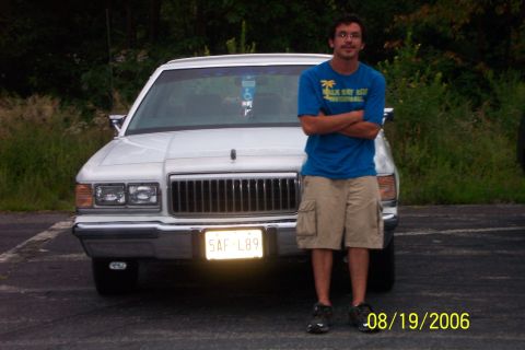 1985 Ford LTD Crown Victoria - A white sedan with a distinctive grille and headlights is shown from the front, with a man standing beside it.