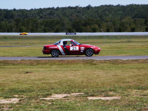 2001 Mercury Grand Marquis - A red Mazda Miata with racing number 21 drives on a track, with another yellow car visible in the distance.