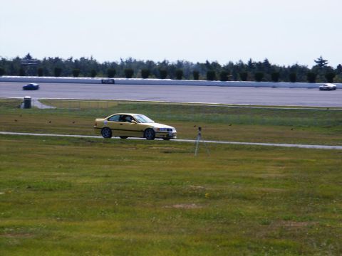 2001 Mercury Grand Marquis - A yellow BMW E36 coupe drives on a racetrack with other vehicles visible.