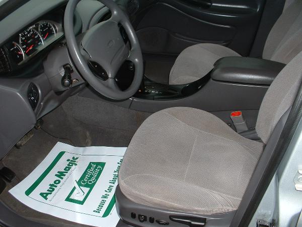 Gray cloth interior of a 1998 Ford Taurus, showing the dashboard, steering wheel, center console, and front seats. A floor mat with "Auto Magic" is visible