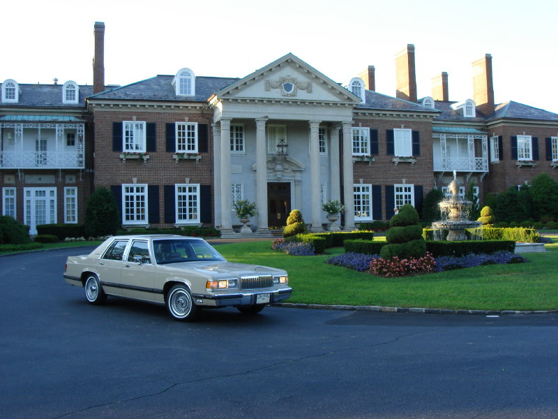 Tan 1990 Mercury Grand Marquis sedan parked on a dark asphalt driveway in front of a large brick mansion with a fountain