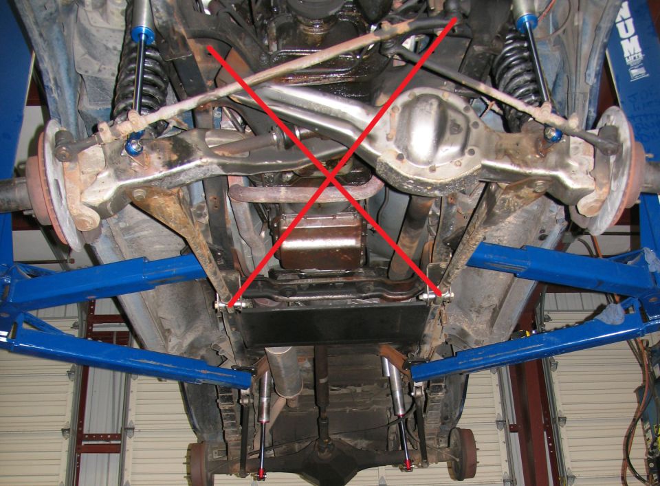 Undercarriage view of a vehicle on a lift, showing suspension and drivetrain components. Red X marks are overlaid