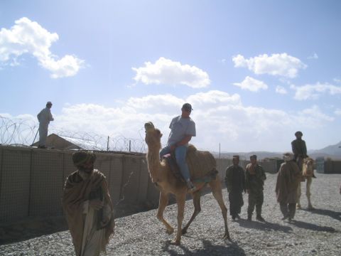 1979 Ford Bronco - A man rides a camel in a dusty area with a fortified wall and barbed wire in the background.