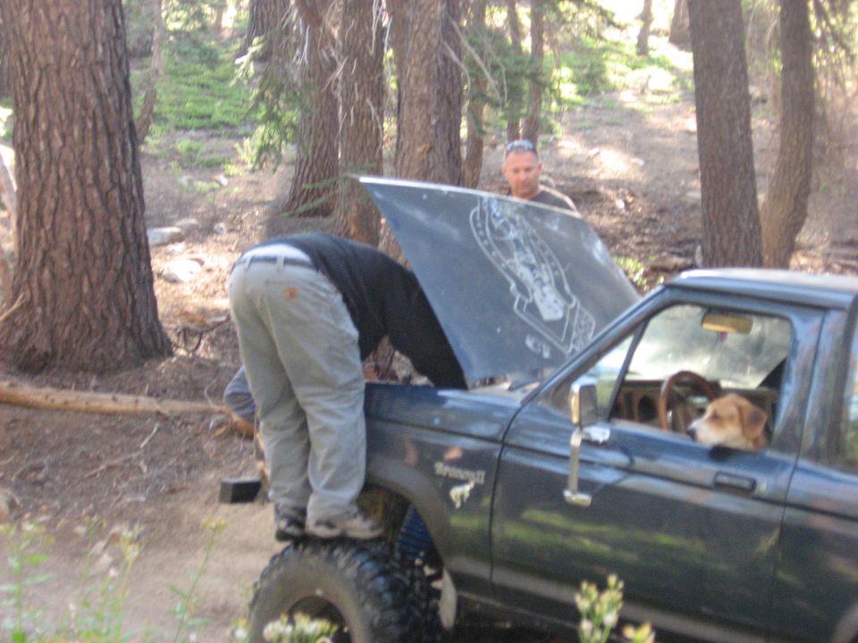 Guy working on a 1986 Ford Bronco II with the hood up, another guy watches, dog in the passenger seat