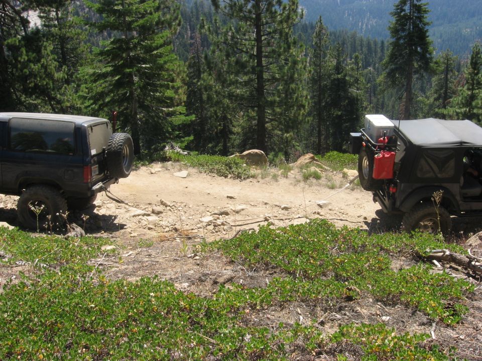 Dark blue SUV with spare tire on back is being pulled by a black Jeep on a rocky, wooded trail