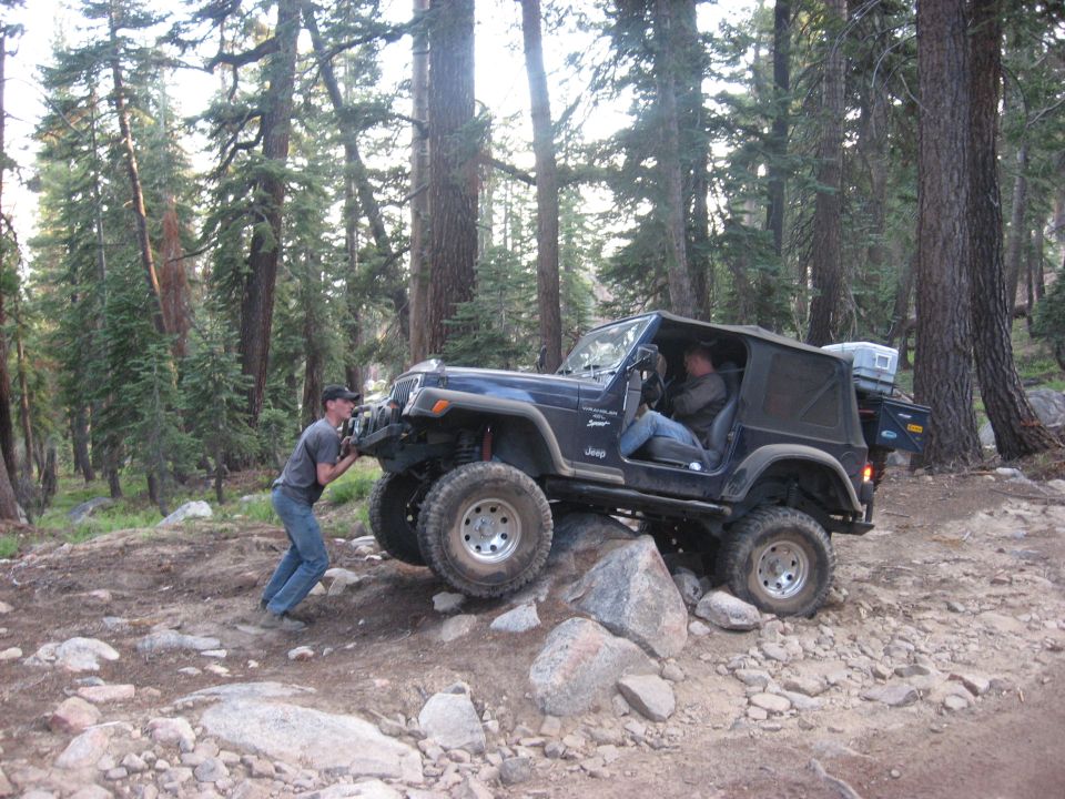 Black Jeep Wrangler crawling over rocks on forest trail with spotter pushing