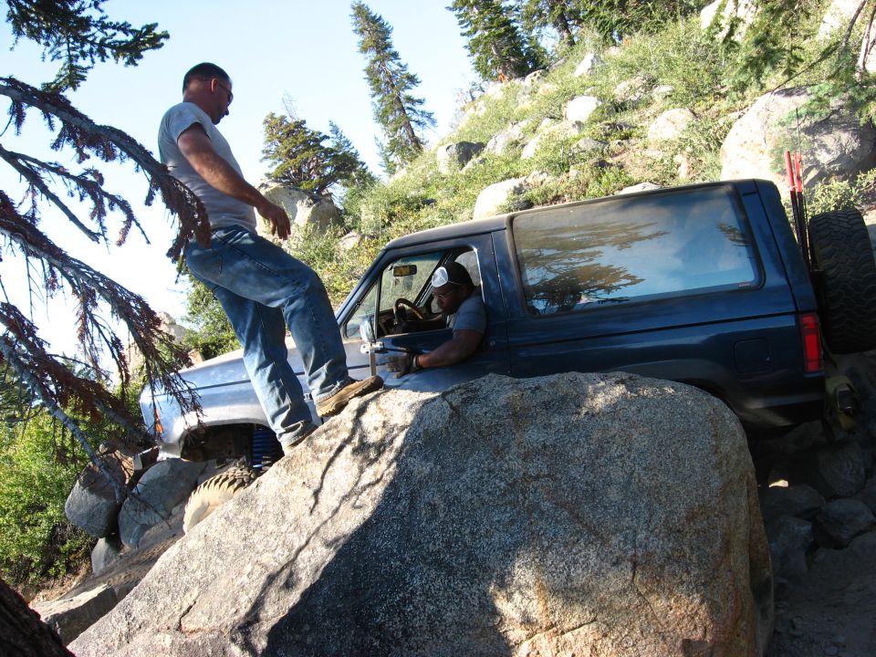 Dark blue Ford Bronco II on a rocky trail, driver visible inside, another vehicle partially seen behind