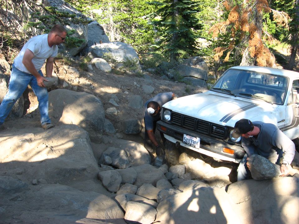 White Toyota pickup truck navigating a rocky trail, people moving rocks